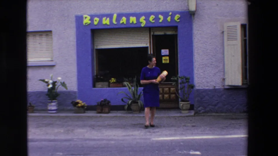 A 1969 Loudes, France home-movie frame shows a woman carrying a baguette near an open boulangerie doorway with storefront lettering and plants along the sidewalk.