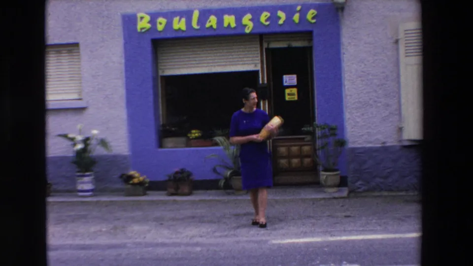 The woman remains outside the Loudes boulangerie in 1969, holding a baguette against a storefront with green lettering, a window display, and potted plants.