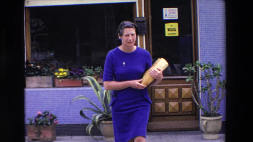 A closer 1969 Loudes, France frame shows the woman facing the camera with a baguette in her arms, standing before the boulangerie window, doorway, and potted plants.