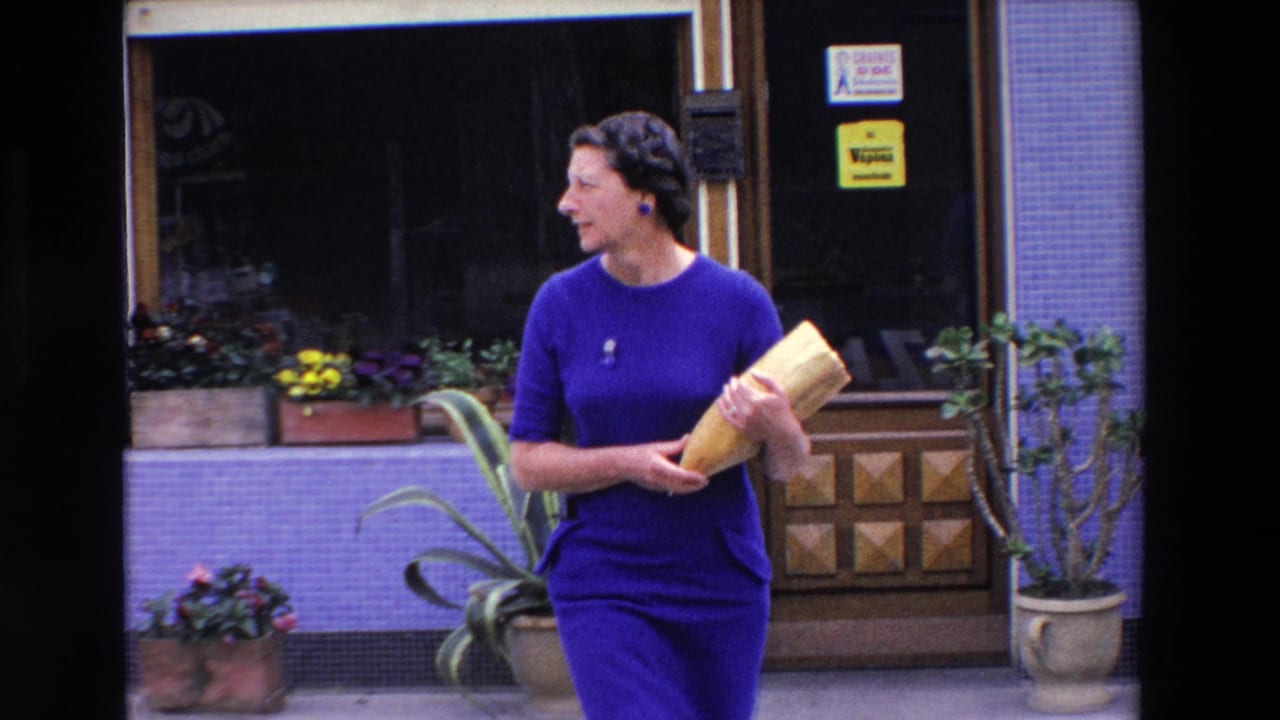 In 1969 France, a woman in a vivid blue dress stands near a shop entrance holding a large cut loaf of bread, with tiled walls and flower boxes behind her.