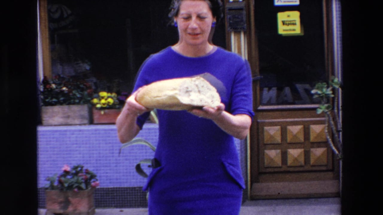 The woman in a blue dress presents the large half loaf toward the camera outside a French shop, with flowers, tiled walls, and signage visible in the background.
