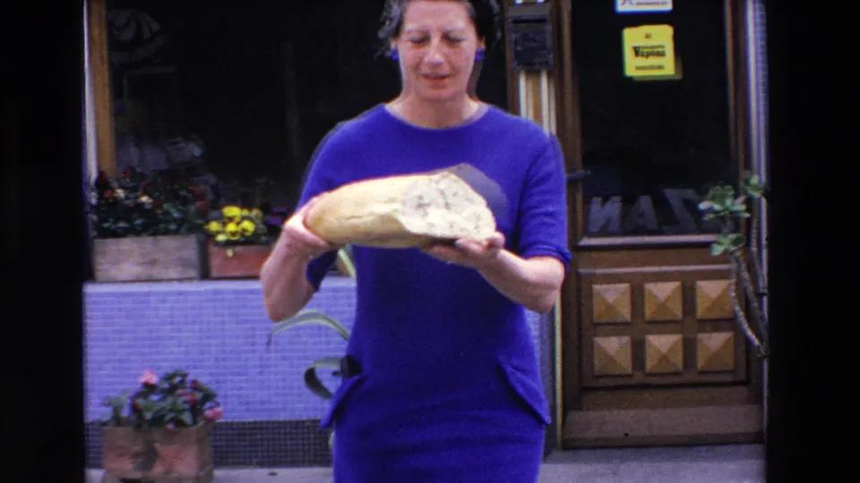 The woman in a blue dress presents the large half loaf toward the camera outside a French shop, with flowers, tiled walls, and signage visible in the background.