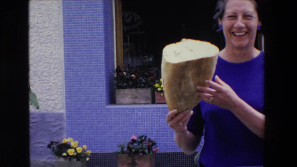 A smiling woman in blue holds the cut face of a large loaf toward the camera in the 1969 France home-movie scene, with a storefront and flowers behind her.
