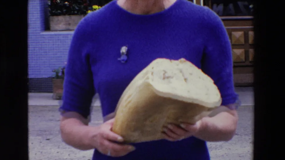 Close framing shows the woman’s blue dress and hands holding the large cut loaf, with the shop facade and doorway partly visible behind her.