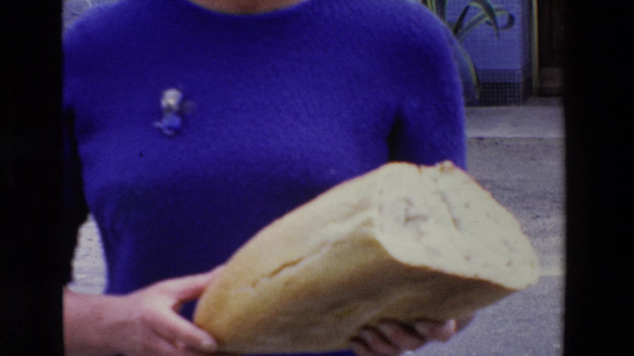 In 1969 home-movie footage from Loudes, France, a woman in a vivid blue dress holds a large loaf of bread close to the camera beside an outdoor entry area.