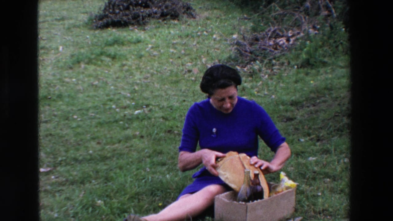 The woman remains seated on the grass in the 1969 France picnic scene, holding the opened paper-wrapped bread over a cardboard box with dark bottles and picnic items.