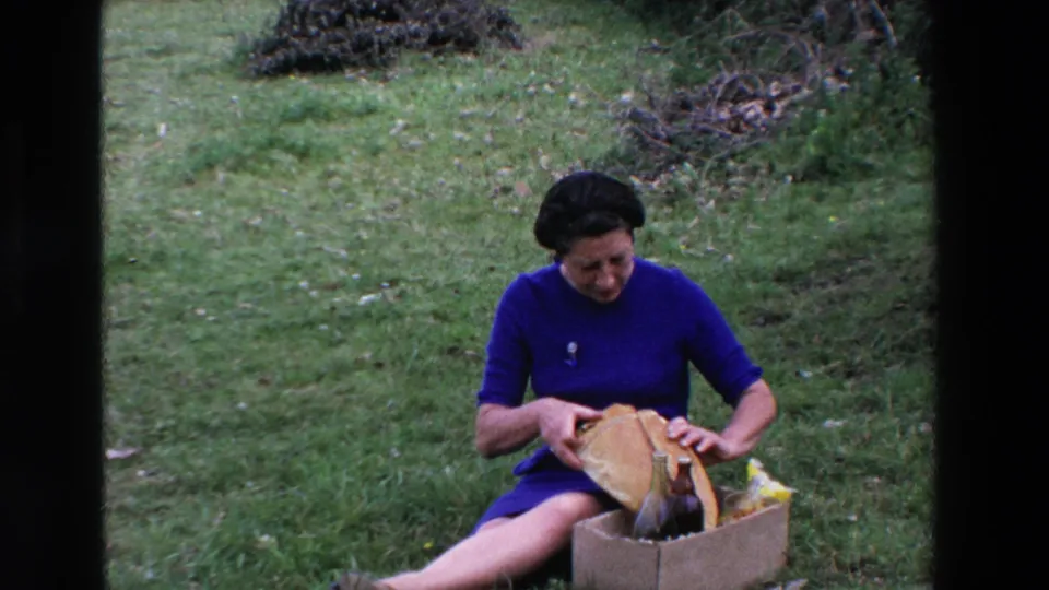 The woman remains seated on the grass in the 1969 France picnic scene, holding the opened paper-wrapped bread over a cardboard box with dark bottles and picnic items.