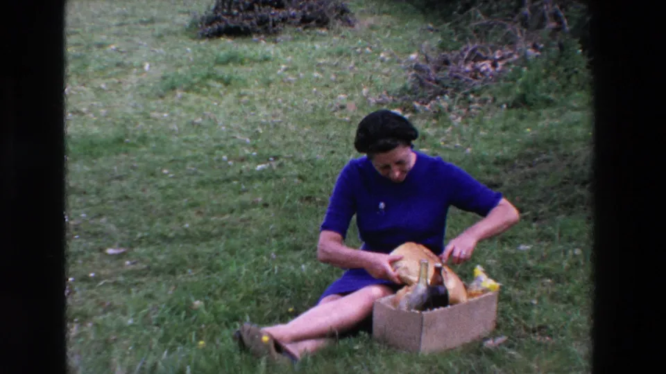 In supported 1969 Loudes, France home-movie footage, a woman in a blue dress sits on grass with a cardboard box of picnic food and bread.