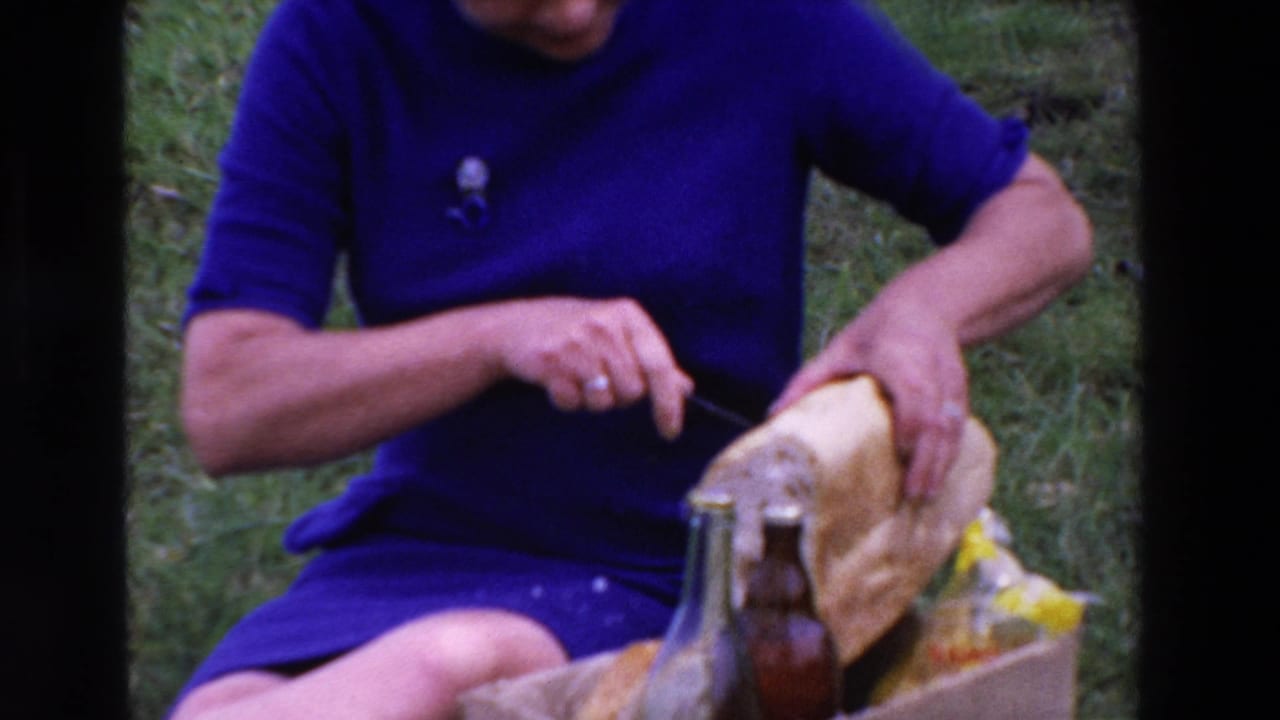Close 8mm frame from the 1969 Loudes, France picnic shows the woman holding bread and cutting it with a knife beside glass bottles and food wrapping.
