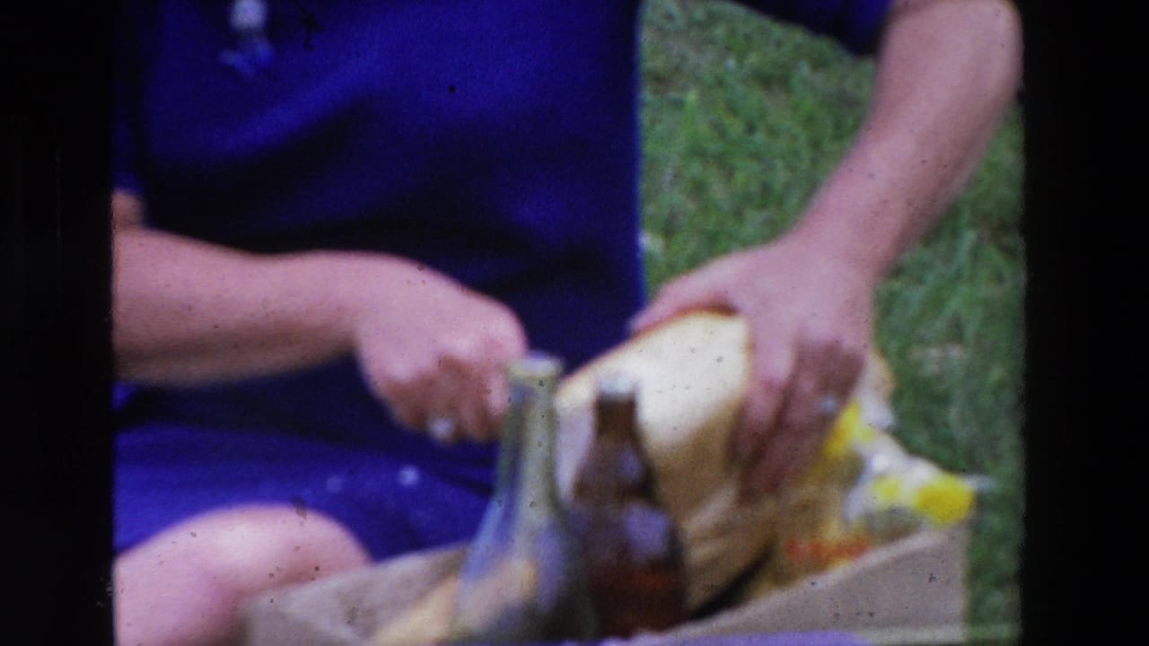 Close 8mm frame of a woman in a blue dress cutting a loaf of bread during a grassy picnic in the 1969 France travel footage.