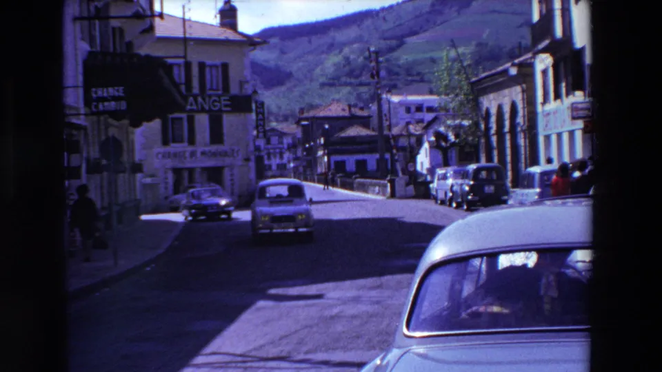 Street view in the 1969 France home movie showing vintage cars moving through a hillside town with visible ANGE and CHANGE DE MONNAIES signage.