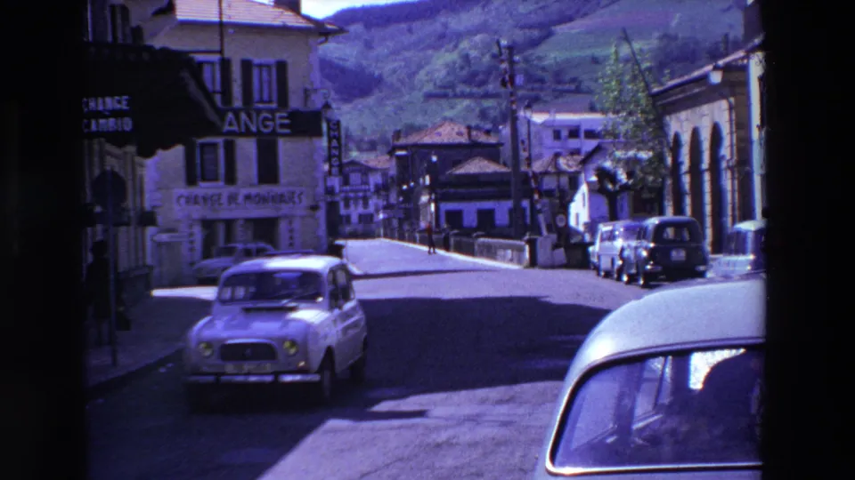 A light-colored vintage sedan drives along a French town street in the 1969 8mm travel footage, with buildings, signs, and hills beyond.