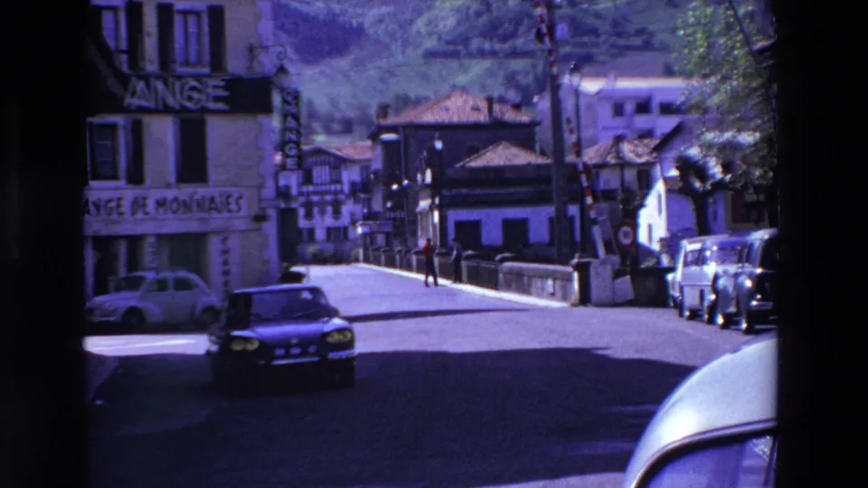 Final street frame from the 1969 France clip, showing a dark car passing through a hillside town area with readable exchange signage and parked vehicles.