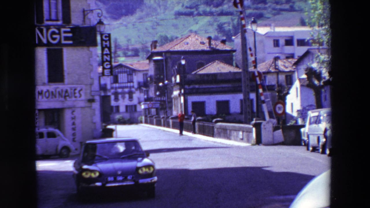 A 1969 Lourdes, France street view shows a dark car driving past a building with "CHANGE" and "MONNAIES" signage, with tiled roofs and a hillside behind.