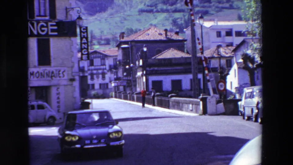 A 1969 Lourdes, France street view shows a dark car driving past a building with "CHANGE" and "MONNAIES" signage, with tiled roofs and a hillside behind.