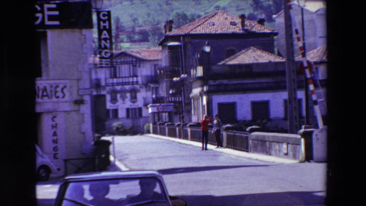 A Lourdes street scene shows the exchange building at left, striped barriers near the roadway, and two men standing on a bridge in bright daylight.