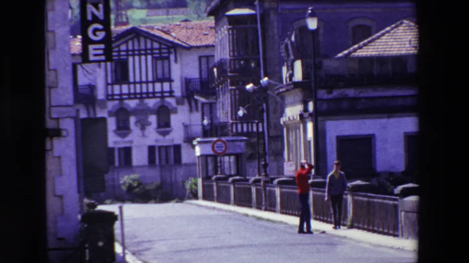 Two men, one wearing a red shirt, stand on a bridge in Lourdes, France, with timber-trimmed facades, street lamps, and exchange signage nearby.