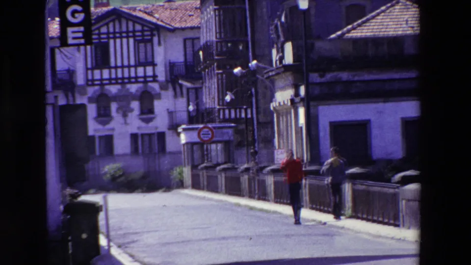 The final frame holds on two men beside the bridge railing in Lourdes, with a street sign, stucco buildings, tiled roofs, and exchange lettering at the edge of frame.