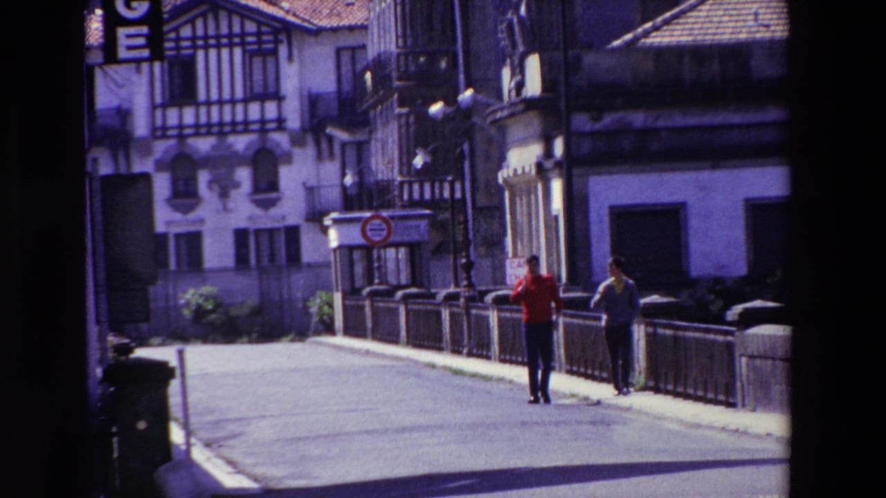 In the supplied 1969 Lourdes, France clip, two adult men walk on a paved street beside a low stone railing, with half-timbered architecture and balconies in the background.