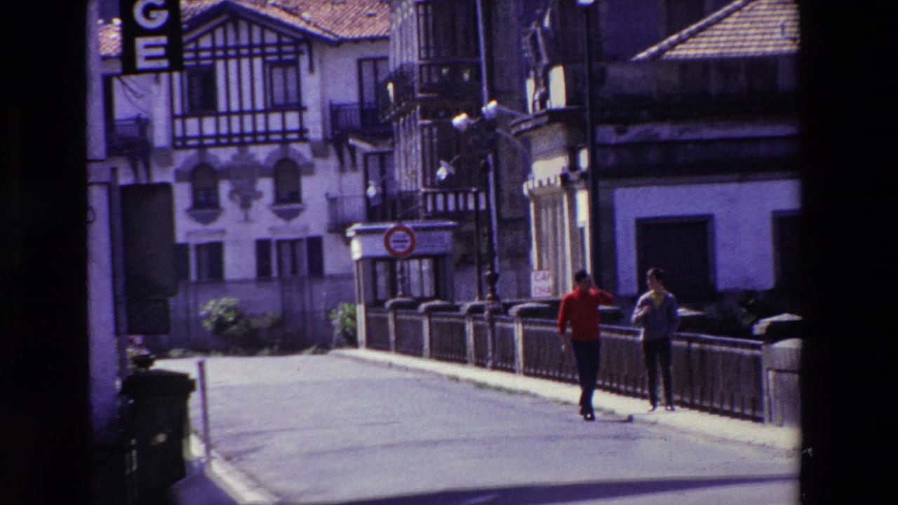 Two adult men continue along the quiet paved street in the 1969 Lourdes footage, passing a low railing with half-timbered and stucco building facades behind them.