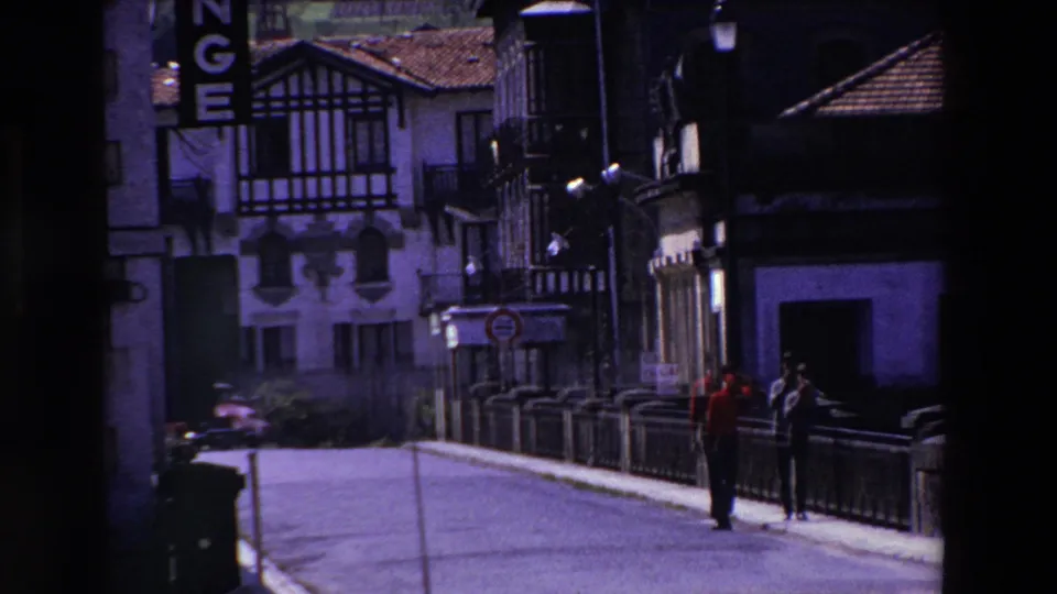 The two adult men appear farther along the street, framed by a low railing, dark shaded facades, a half-timbered building, and a vertical sign at the edge of the frame.