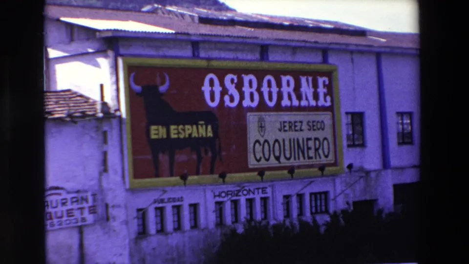1969 Lourdes, France 8mm frame showing the Osborne sign on the building, with the black bull graphic, EN ESPAÑA text, and white Coquinero panel visible above small windows.
