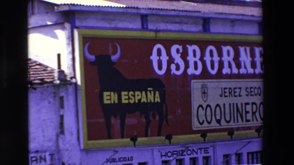 1969 8mm frame showing a large red Spanish-language Osborne billboard with a black bull silhouette, yellow EN ESPAÑA text, and a smaller Jerez Seco Coquinero panel beside building edges.