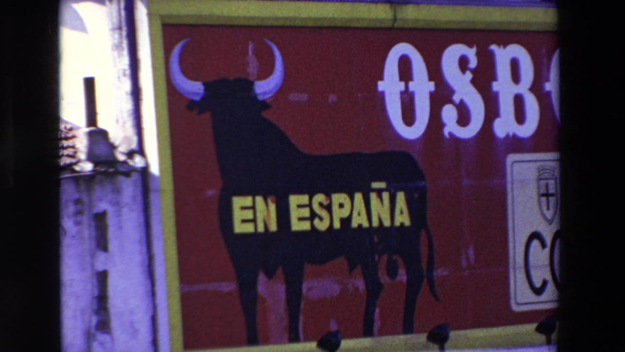 1969 8mm frame with a closer view of the red Osborne sign, black bull silhouette, yellow EN ESPAÑA lettering, partial white brand lettering, and surrounding building edges.