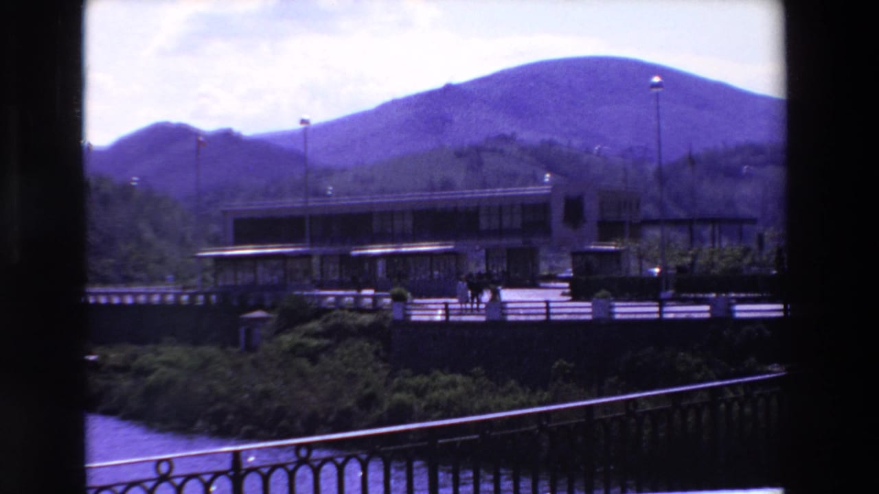 1969 home-movie view in France of a low modernist building at the base of mountains, framed by dark vertical edges with a railing and water below.