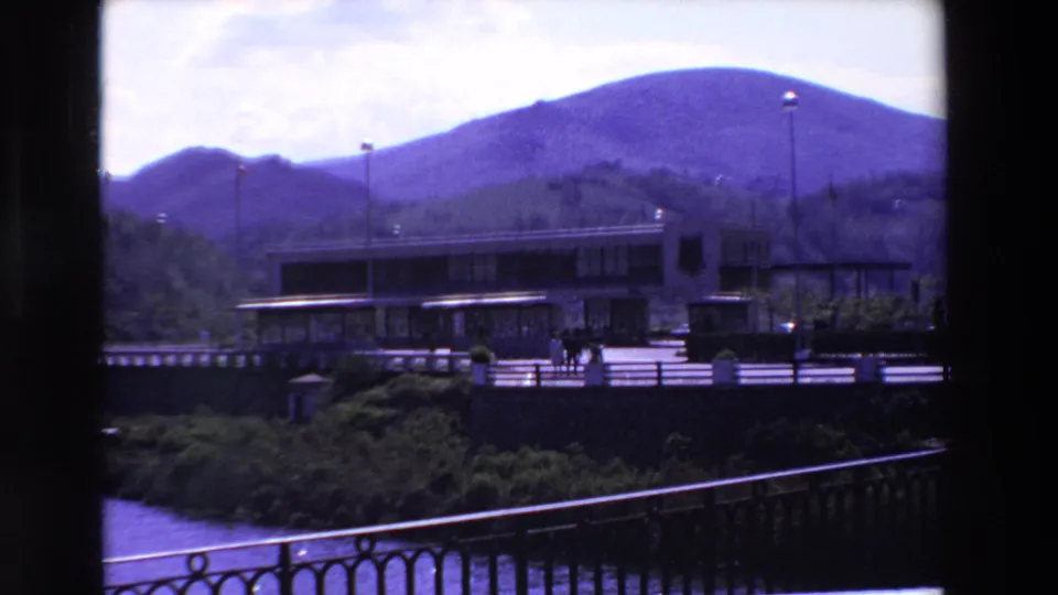 1969 home-movie view in France of a low modernist building at the base of mountains, framed by dark vertical edges with a railing and water below.