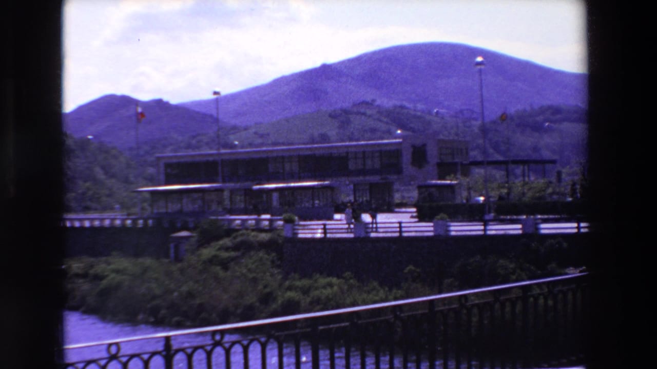 Low building and flagpoles below purple-blue mountains with pedestrians on the foreground walkway.