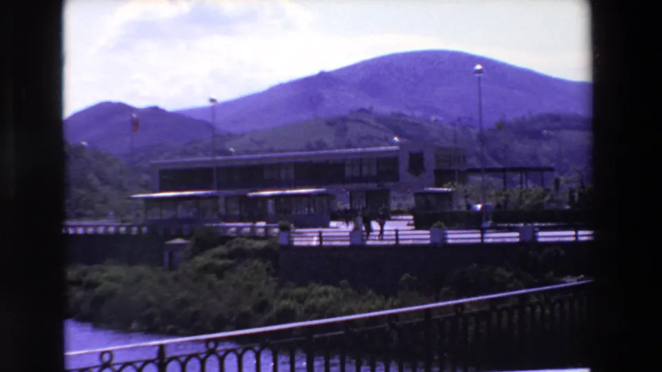 1969 home-movie frame in France with a modern riverside building, mountain range, flagpoles, small pedestrians, foliage, and ornate railing in view.