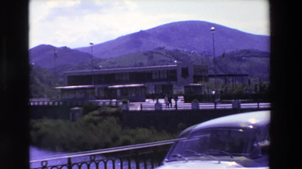 Final 1969 France frame showing a rounded car passing in the foreground, with a modern building, pedestrians, flagpoles, railing, foliage, and mountains behind.