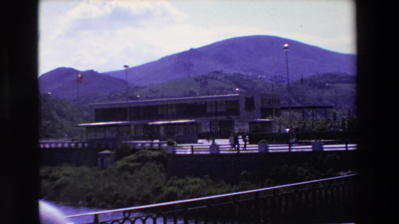 1969 8mm home-movie frame from Loudes, France, showing a modern low building by water, small pedestrians, flagpoles, railings, and purple-hued mountains behind.