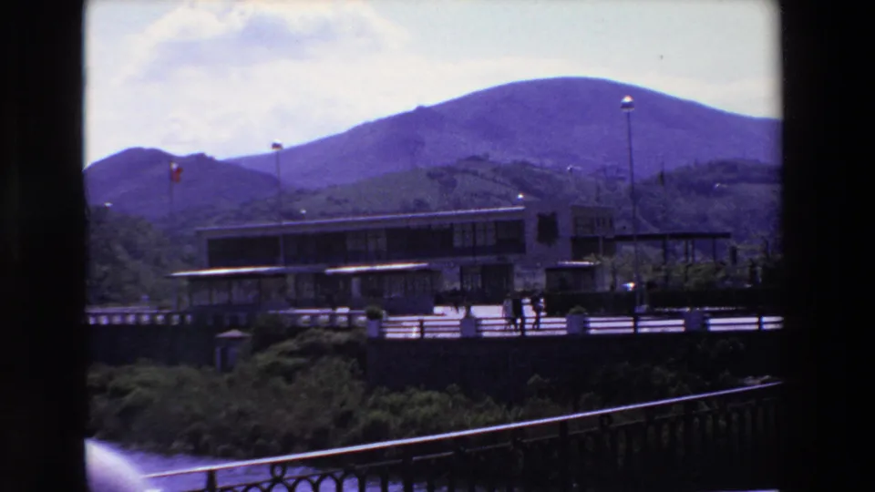 1969 8mm home-movie frame from Loudes, France, showing a modern low building by water, small pedestrians, flagpoles, railings, and purple-hued mountains behind.