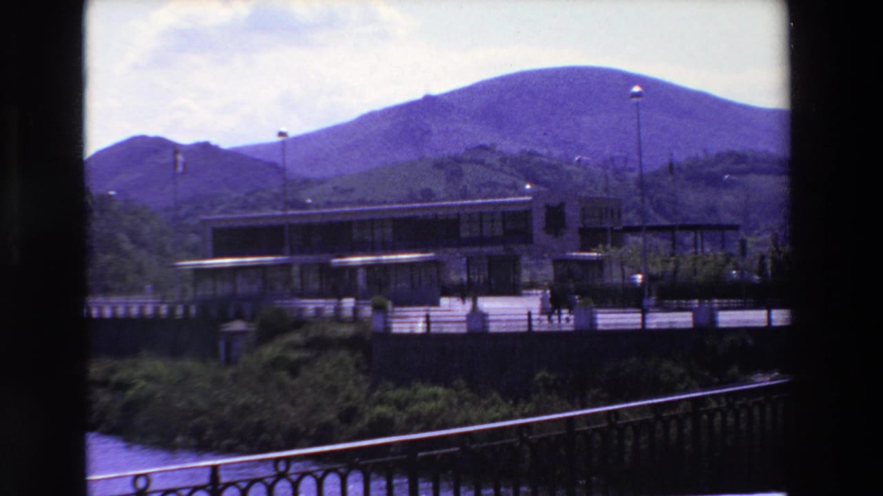 1969 8mm frame from Loudes, France, with a modern riverside building, several small walking figures, flagpoles, foreground railing, and mountains under a bright sky.