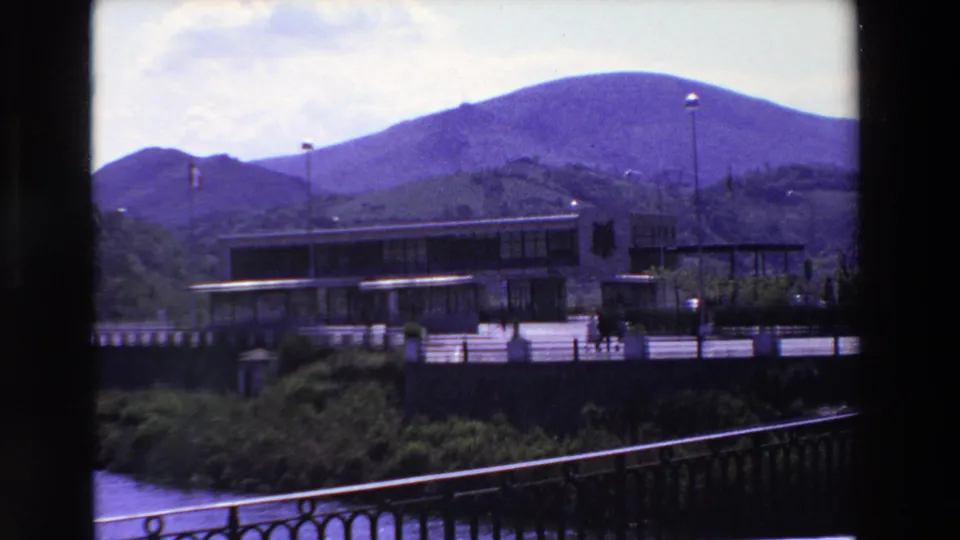 1969 8mm frame from Loudes, France, with a modern riverside building, several small walking figures, flagpoles, foreground railing, and mountains under a bright sky.