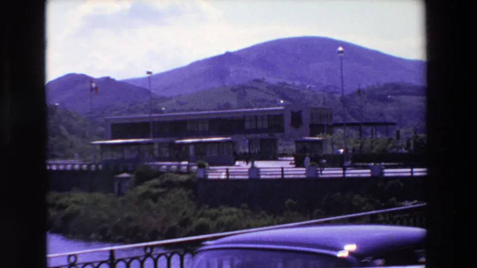1969 8mm home-movie frame from Loudes, France, showing pedestrians near a modern building by water, a dark railing, mountain slopes, and a partial classic car roof at the lower edge.