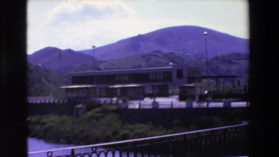 Final 1969 8mm frame from Loudes, France, with pedestrians crossing near a modern building, flagpoles, a foreground railing, water below, and purple-tinted mountains in the background.