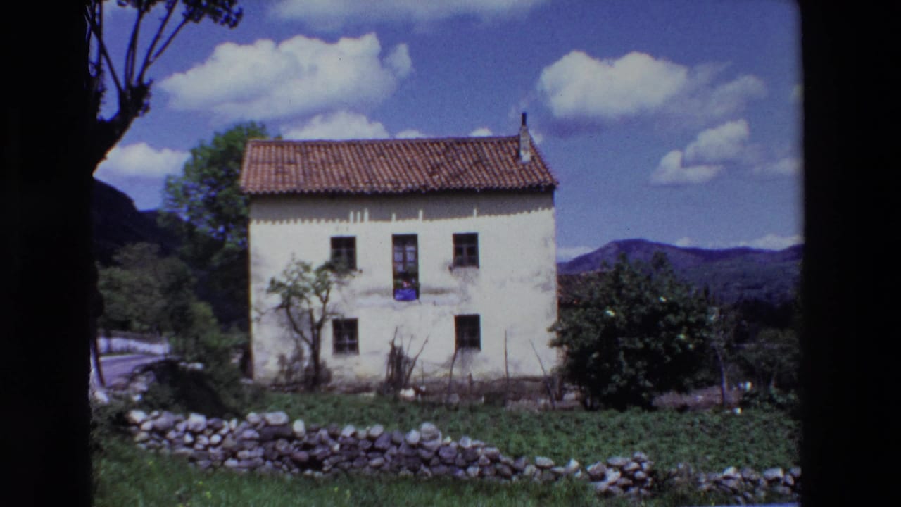 1969 Lourdes, France 8mm frame showing a white two-story house with a red tile roof, dark windows, a low stone wall, shrubs, distant hills, and clouds overhead.