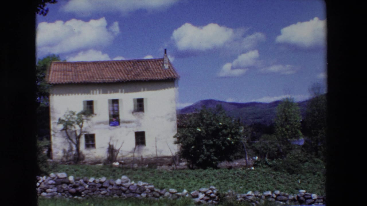 1969 Lourdes, France frame with the white house centered, a red tile roof, visible windows, garden vegetation, a low stone wall, rolling hills, and scattered clouds.