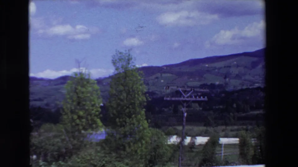 1969 Lourdes, France frame looking away from the house across trees, green valley vegetation, a utility pole, distant hills, and a bright sky with scattered clouds.