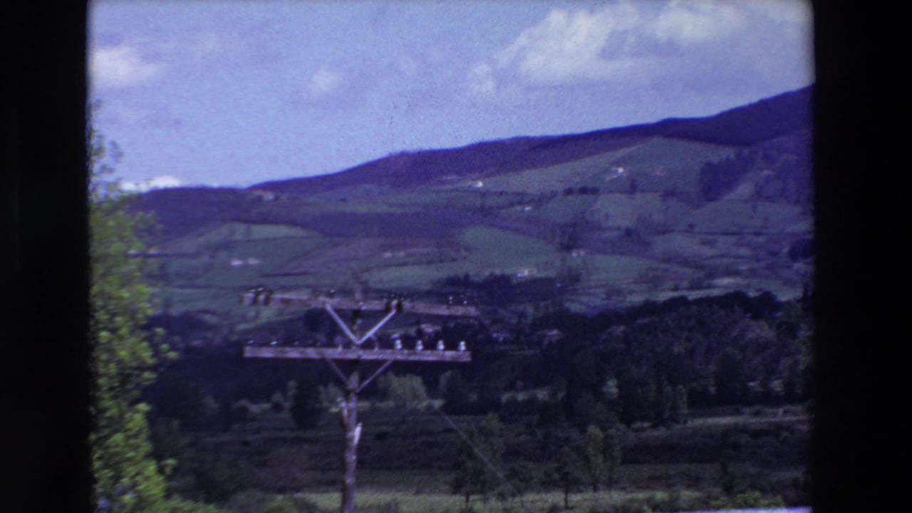 1969 8mm frame near Loudes, France, showing rolling green fields below a wooded ridge with a wooden utility pole and wires in the foreground.