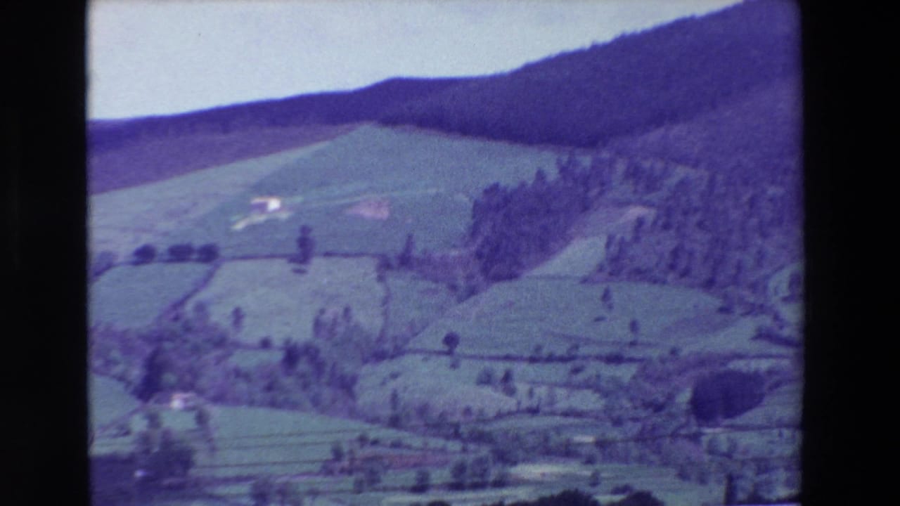 1969 8mm frame near Loudes, France, showing patchwork meadows, tree clusters, and small pale hillside buildings under a bright sky.