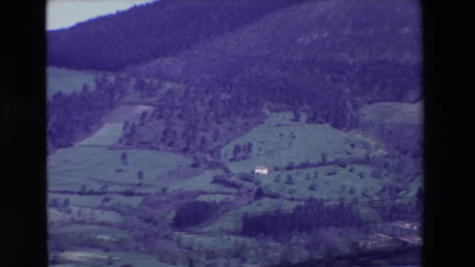 1969 8mm frame near Loudes, France, with soft-focus meadows, tree lines, a wooded mountain slope, and small white structures set into the landscape.