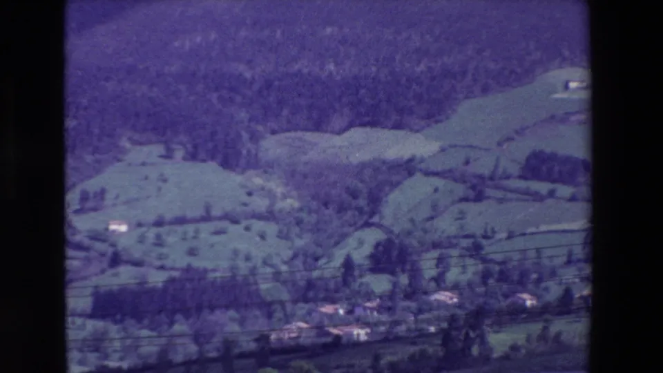 1969 8mm frame near Loudes, France, showing a wide rural valley with green fields, wooded slopes, overhead wires, and a small cluster of rooftops near the lower edge.