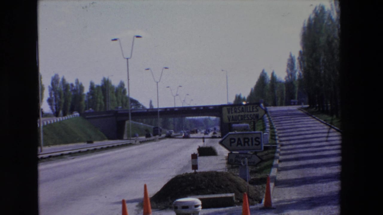 1969 Paris-region highway entrance with directional signs for Paris, Versailles, and Vaucresson, cars on the road, and an overpass ahead.