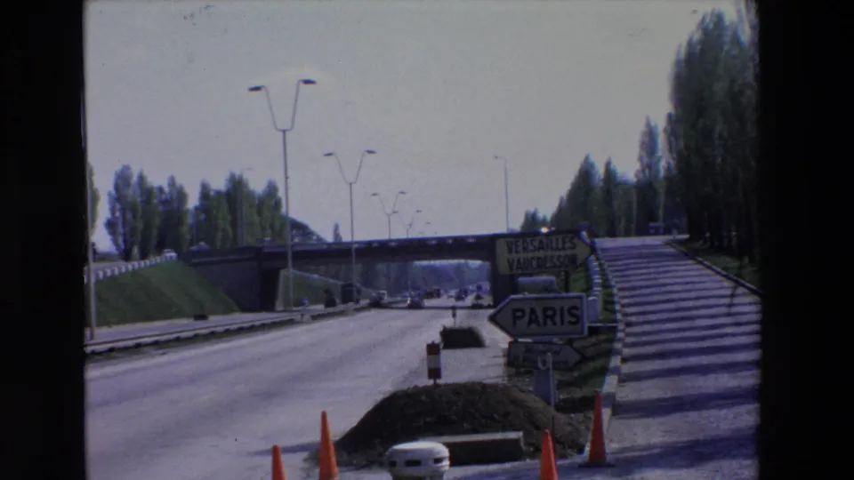 1969 Paris-region highway entrance with directional signs for Paris, Versailles, and Vaucresson, cars on the road, and an overpass ahead.