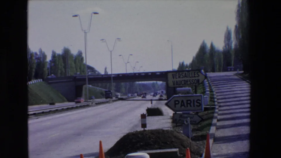 Paris-area roadside view with highway lanes, cars, a pedestrian-style stair embankment, and signs indicating Paris, Versailles, and Vaucresson.