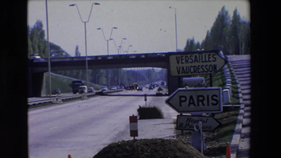 Closer 1969 view of Paris and Versailles-Vaucresson directional signs at a highway entrance, with cars, an overpass, cones, and embankment stairs visible.